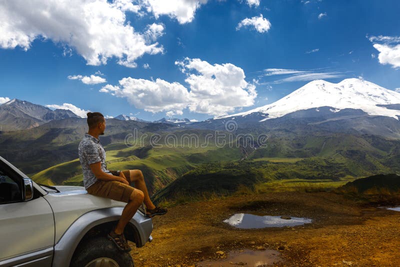 De jonge Reizigersmens zit op Auto en geniet van Mening van Bergen in de Zomer Elbrusgebied, de Noord-Kaukasus, Rusland royalty-vrije stock afbeeldingen De jonge Reizigersmens zit op Auto en geniet van Mening van Bergen in de Zomer Elbrusgebied, de Noord-Kaukasus, Rusland royalty-vrije stock afbeeldingen