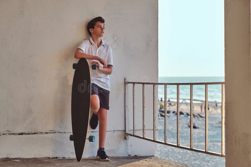 De jonge schaatserjongen kleedde zich in t-shirt en borrels die op een muur leunen en houdt een skateboard tegen de achtergrond v stock afbeeldingen De jonge schaatserjongen kleedde zich in t-shirt en borrels die op een muur leunen en houdt een skateboard tegen de achtergrond v stock afbeeldingen
