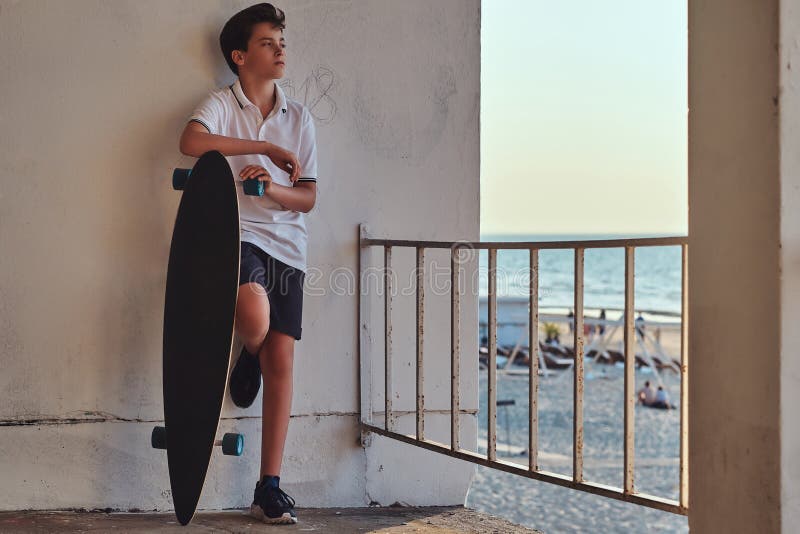 De jonge schaatserjongen kleedde zich in t-shirt en borrels die op een muur leunen en houdt een skateboard tegen de achtergrond v stock fotografie De jonge schaatserjongen kleedde zich in t-shirt en borrels die op een muur leunen en houdt een skateboard tegen de achtergrond v stock fotografie