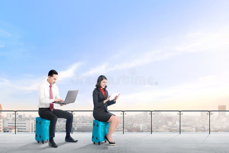 De jonge twee Aziatische bedrijfsmensen die op blauwe koffer zitten houden laptop en de tablet werkend aan modern terras stock foto's De jonge twee Aziatische bedrijfsmensen die op blauwe koffer zitten houden laptop en de tablet werkend aan modern terras stock foto's