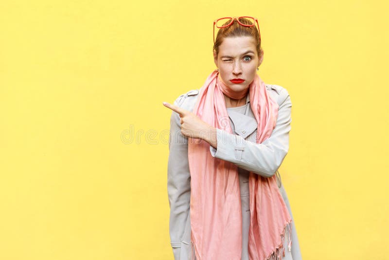 Young adult woman pointing copy space and wink. Seriously looking at camera and pointing away while standing isolated on yellow background. Studio shot. Young adult woman pointing copy space and wink. Seriously looking at camera and pointing away while standing isolated on yellow background. Studio shot