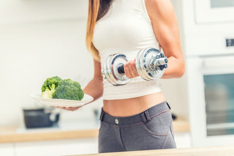 De jonge vrouw met sporten komt in de domoren van de keukenholding in haar handen en een plaat van broccoli voor stock foto De jonge vrouw met sporten komt in de domoren van de keukenholding in haar handen en een plaat van broccoli voor stock foto