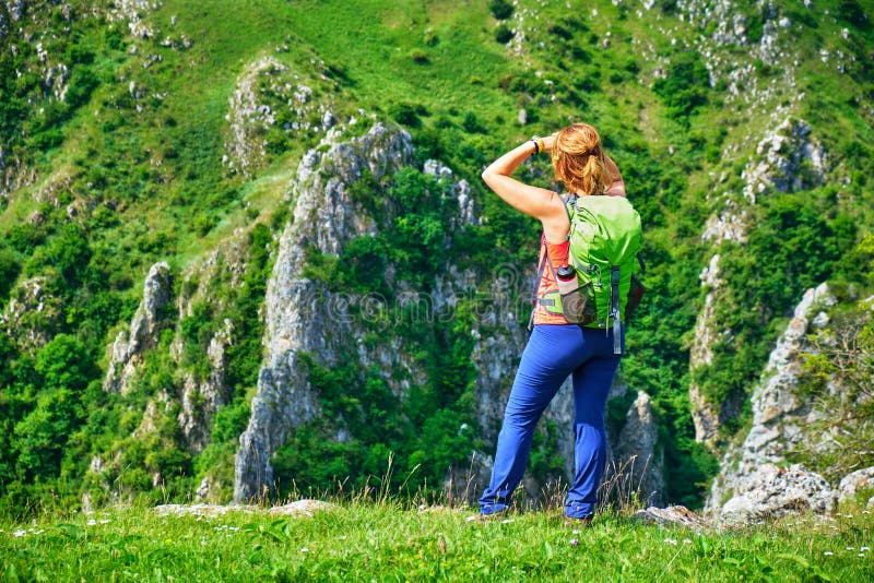 De jonge vrouwenwandelaar met een groene rugzak bevindt zich op een grasrand en kijkt naar de massieve rotsmuren in Tureni-kloof royalty-vrije stock fotografie De jonge vrouwenwandelaar met een groene rugzak bevindt zich op een grasrand en kijkt naar de massieve rotsmuren in Tureni-kloof royalty-vrije stock fotografie