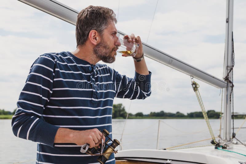 De kapitein met baard en verrekijkers drinkt whisky op jacht stock foto's De kapitein met baard en verrekijkers drinkt whisky op jacht stock foto's