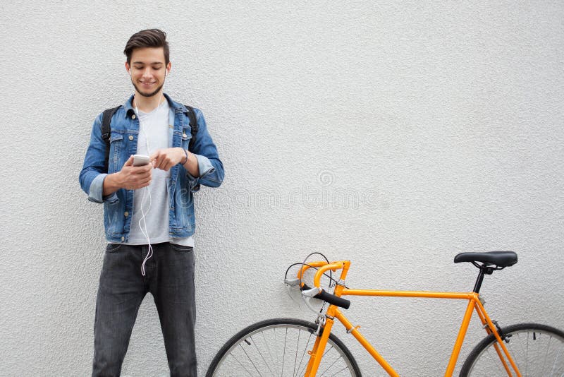 De kerel in een blauw denimjasje die zich op muurachtergrond bevinden jonge mens dichtbij oranje fiets Glimlachende student met z royalty-vrije stock foto De kerel in een blauw denimjasje die zich op muurachtergrond bevinden jonge mens dichtbij oranje fiets Glimlachende student met z royalty-vrije stock foto