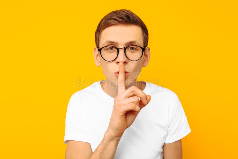 The guy in the white T-shirt in round transparent glasses says: Shhhh, shows a silent gesture, keeps something secret on a yellow background. The guy in the white T-shirt in round transparent glasses says: Shhhh, shows a silent gesture, keeps something secret on a yellow background