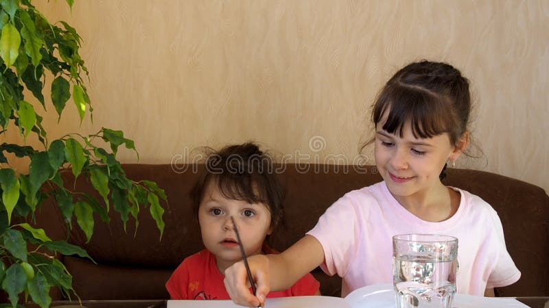 De kinderen trekken met kleuren Twee meisjes trekken huizen op de lijst met verven Een borstel wordt gewassen in een duidelijk gl stock footage De kinderen trekken met kleuren Twee meisjes trekken huizen op de lijst met verven Een borstel wordt gewassen in een duidelijk gl stock footage