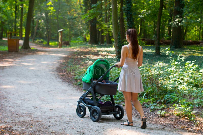 De kinderwagen die terug mening lopen, brengt achtermening van aantrekkelijke vrouw die in modieuze de zomerkleding met haar baby royalty-vrije stock foto's De kinderwagen die terug mening lopen, brengt achtermening van aantrekkelijke vrouw die in modieuze de zomerkleding met haar baby royalty-vrije stock foto's