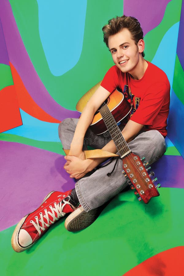 De knappe gelukkige jongen speelt op akoestische gitaar op gekleurde bedelaars royalty-vrije stock foto De knappe gelukkige jongen speelt op akoestische gitaar op gekleurde bedelaars royalty-vrije stock foto