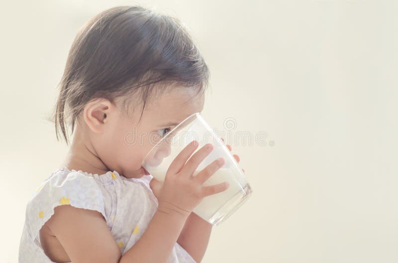 De leuke Aziatische consumptiemelk van het peutermeisje van groot glas op witte bedelaars stock foto De leuke Aziatische consumptiemelk van het peutermeisje van groot glas op witte bedelaars stock foto