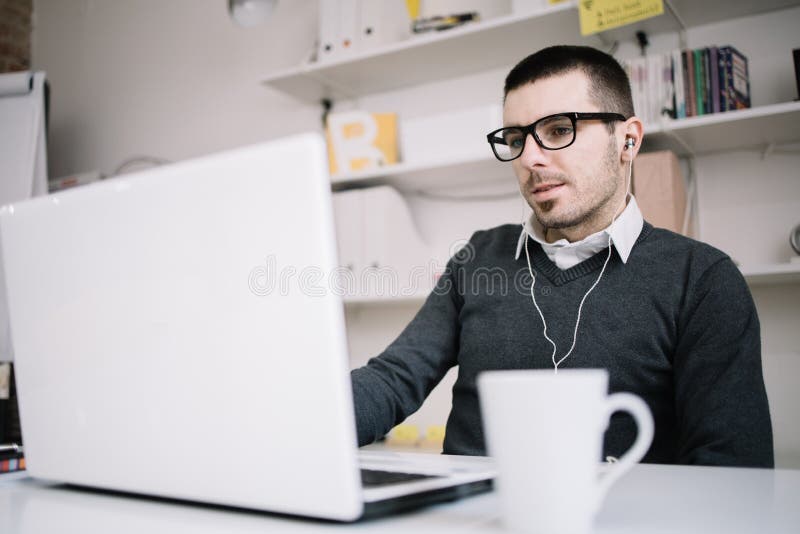 Man communicates with partners online using laptop. Office guy takes part in online meeting while sitting in office and wearing headphones and glasses. Man communicates with partners online using laptop. Office guy takes part in online meeting while sitting in office and wearing headphones and glasses.