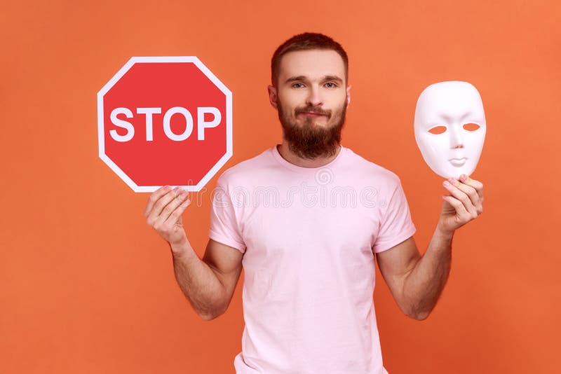 Portrait of puzzled confused bearded man holding white mask with unknown face and red traffic sign, looking at camera, wearing pink T-shirt. Indoor studio shot isolated on orange background. Portrait of puzzled confused bearded man holding white mask with unknown face and red traffic sign, looking at camera, wearing pink T-shirt. Indoor studio shot isolated on orange background.
