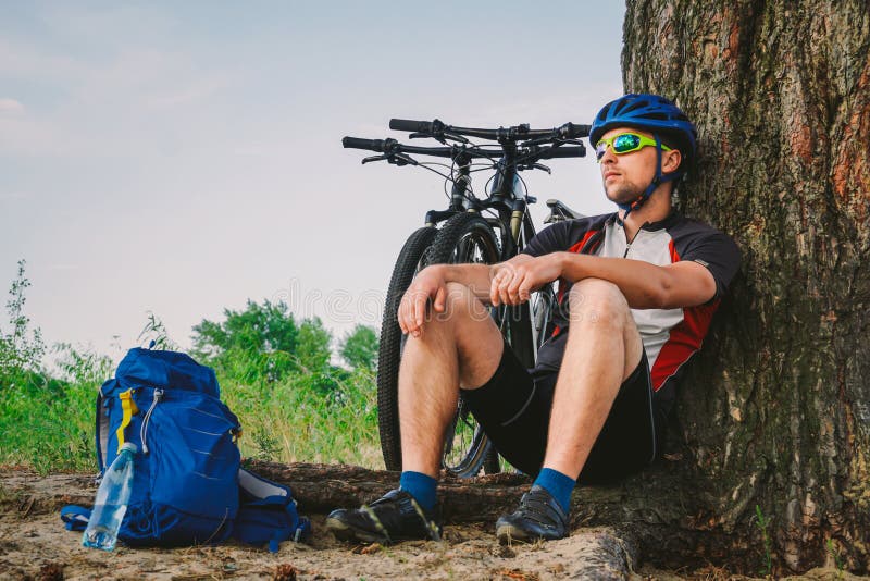 De mannelijke bergfietser die op fietsrit rusten, die op grond onder een boom met zijn bergfiets zitten, bevindt zich naast hem, royalty-vrije stock fotografie De mannelijke bergfietser die op fietsrit rusten, die op grond onder een boom met zijn bergfiets zitten, bevindt zich naast hem, royalty-vrije stock fotografie
