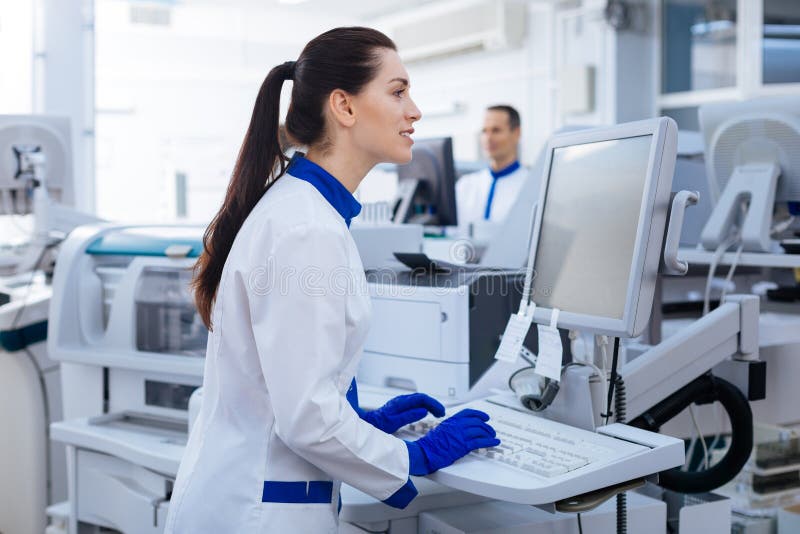 Nice touch. Energetic positive female laboratory assistant looking at the screen, typing while standing near the medical equipment. Nice touch. Energetic positive female laboratory assistant looking at the screen, typing while standing near the medical equipment