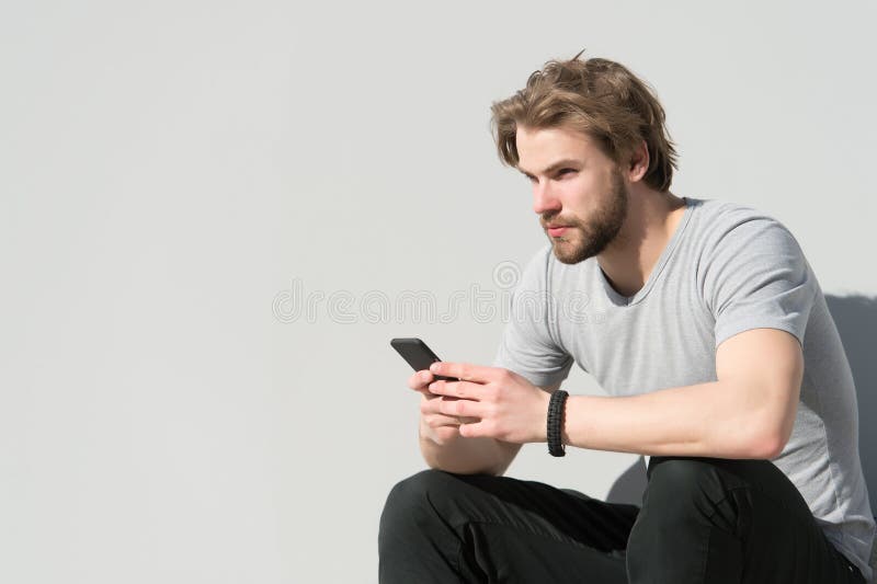 De mens zit met smartphone op grijze muur, nieuwe technologie Macho met baard, blond haar in t-shirt op zonnige dag, manier Nieuw royalty-vrije stock foto's De mens zit met smartphone op grijze muur, nieuwe technologie Macho met baard, blond haar in t-shirt op zonnige dag, manier Nieuw royalty-vrije stock foto's
