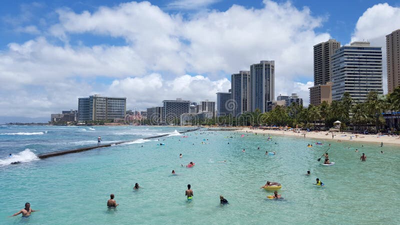 WAIKIKI - July 17, 2016: People play in the protected water and hang out on the beach in world famous tourist area Waikiki on a beautiful day with hotels in the distance. July 3, 2016 in Waikiki, Hawaii. WAIKIKI - July 17, 2016: People play in the protected water and hang out on the beach in world famous tourist area Waikiki on a beautiful day with hotels in the distance. July 3, 2016 in Waikiki, Hawaii.