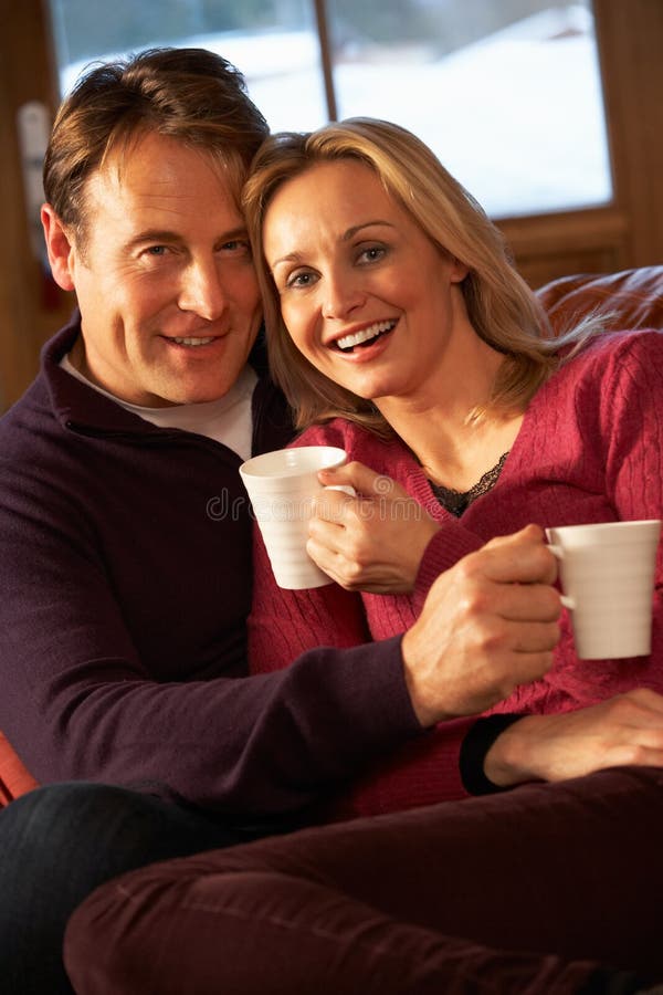 Middle Aged Couple Sitting On Sofa With Hot Drinks In Chalet. Middle Aged Couple Sitting On Sofa With Hot Drinks In Chalet