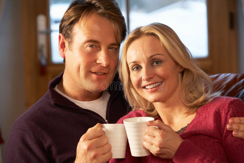 Middle Aged Couple Sitting On Sofa With Hot Drinks In Chalet On Ski Holiday Smiling At Camera. Middle Aged Couple Sitting On Sofa With Hot Drinks In Chalet On Ski Holiday Smiling At Camera