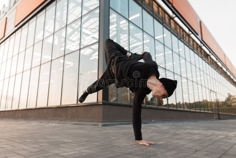 De modieuze jonge hipstermens in modieuze zwarte kleren in zonnebril in een gebreide uitstekende hoed danst in de stad stock afbeeldingen De modieuze jonge hipstermens in modieuze zwarte kleren in zonnebril in een gebreide uitstekende hoed danst in de stad stock afbeeldingen