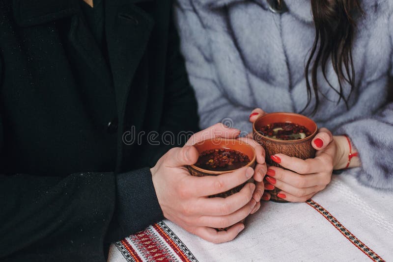 Stylish couple holding cups with hot tea in hands on wooden porch in winter snowy mountains. Happy romantic family with drinks. Holiday getaway together. Romantic moments. Stylish couple holding cups with hot tea in hands on wooden porch in winter snowy mountains. Happy romantic family with drinks. Holiday getaway together. Romantic moments