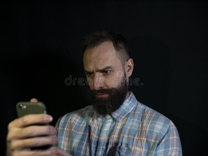 De modieuze mens met een baard en een snor bekijkt een mobiele telefoon met een ernstige gelaatsuitdrukking op een zwarte achterg royalty-vrije stock afbeelding De modieuze mens met een baard en een snor bekijkt een mobiele telefoon met een ernstige gelaatsuitdrukking op een zwarte achterg royalty-vrije stock afbeelding