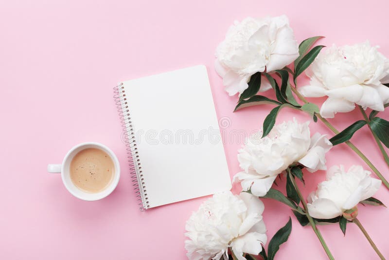 De mok van de ochtendkoffie voor ontbijt, het lege notitieboekje en de witte pioenbloemen op roze de bovenkantmening van de paste