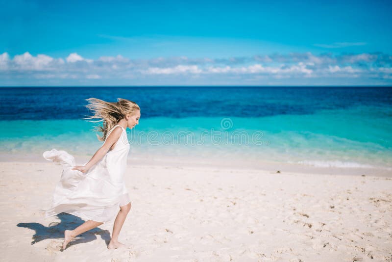 Beautiful blonde long hair bride in long white dress running on the white sand beach. Tropical turquois sea on the background. Beautiful blonde long hair bride in long white dress running on the white sand beach. Tropical turquois sea on the background