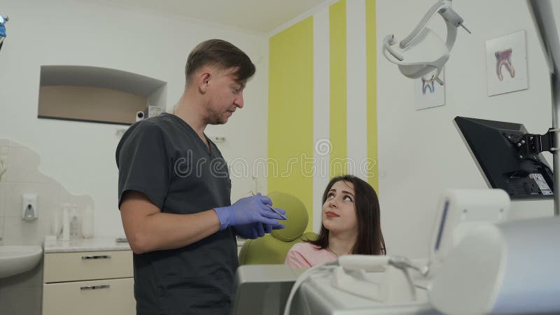 De mooie jonge patiënt glimlacht terwijl het zitten als voorzitter bij de tandarts die van tandartsHandsome kunstmatige kaak tone