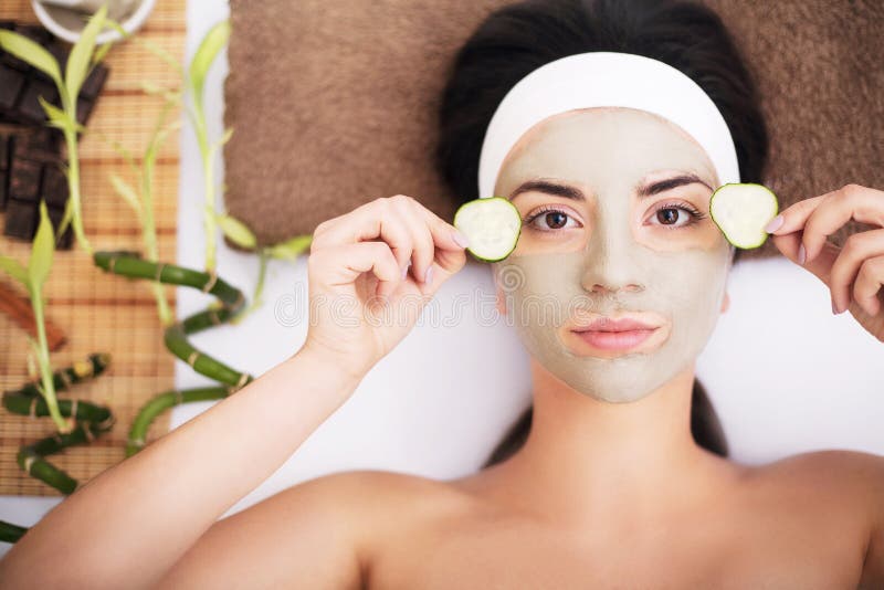 Beautiful young woman is getting facial clay mask at spa, lying with cucumbers on eyes. Beautiful young woman is getting facial clay mask at spa, lying with cucumbers on eyes.