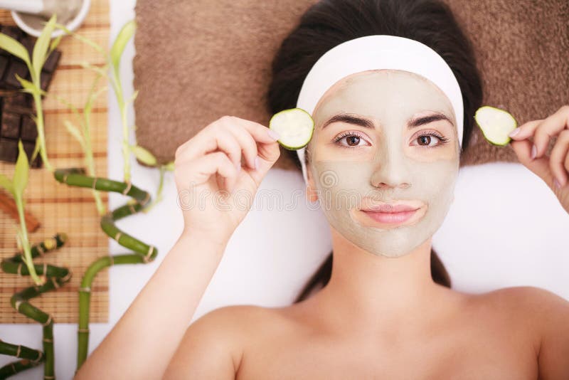 Beautiful young woman is getting facial clay mask at spa, lying with cucumbers on eyes. Beautiful young woman is getting facial clay mask at spa, lying with cucumbers on eyes.