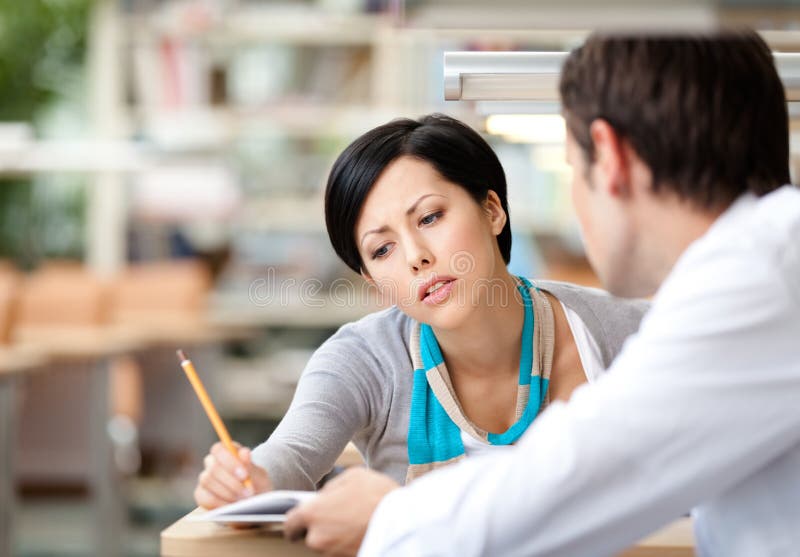 Pretty women communicates with young men showing her something in the book sitting at the desk at the library. Pretty women communicates with young men showing her something in the book sitting at the desk at the library