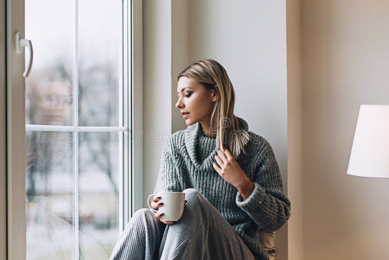 De mooie witte modieuze vrouw in comfortabele Skandinavische interrior zit thuis dichtbij het grote venster, portret van mooi stock afbeeldingen De mooie witte modieuze vrouw in comfortabele Skandinavische interrior zit thuis dichtbij het grote venster, portret van mooi stock afbeeldingen