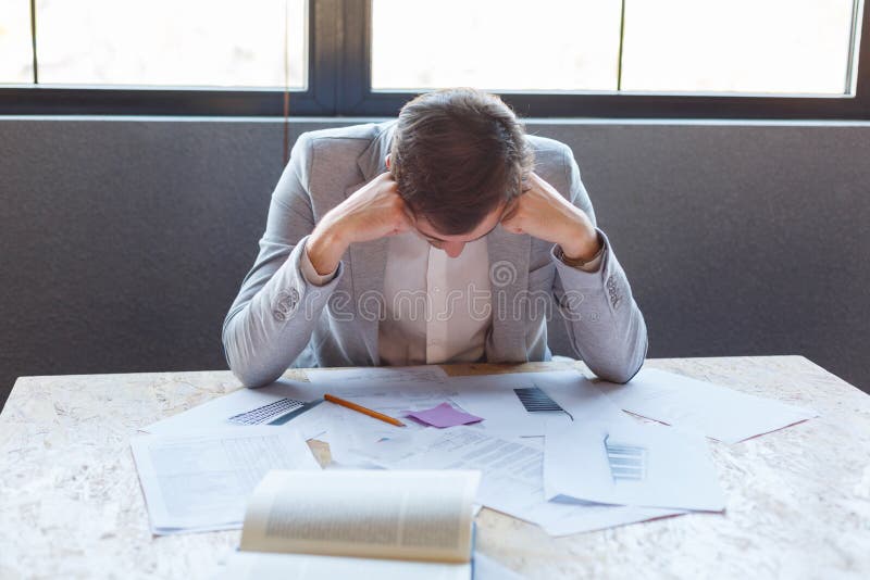 De ongelukkige man zit bij het bureau met documenten, gek overhellend zijn hoofd Binnen in het bureau royalty-vrije stock foto's De ongelukkige man zit bij het bureau met documenten, gek overhellend zijn hoofd Binnen in het bureau royalty-vrije stock foto's