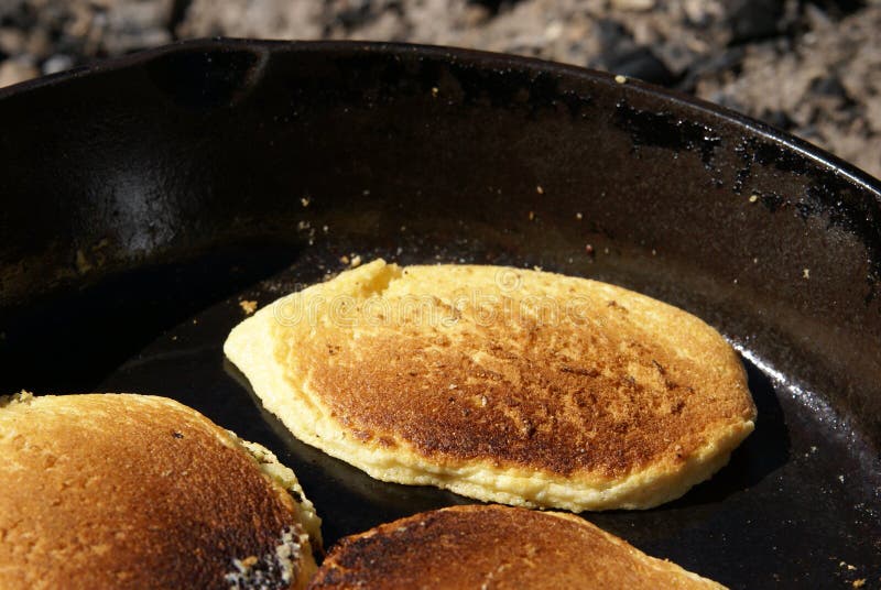 Cornmeal pancakes cooking in a cast iron skillet over an open fire outdoors. Close up view of cakes. Cornmeal pancakes cooking in a cast iron skillet over an open fire outdoors. Close up view of cakes