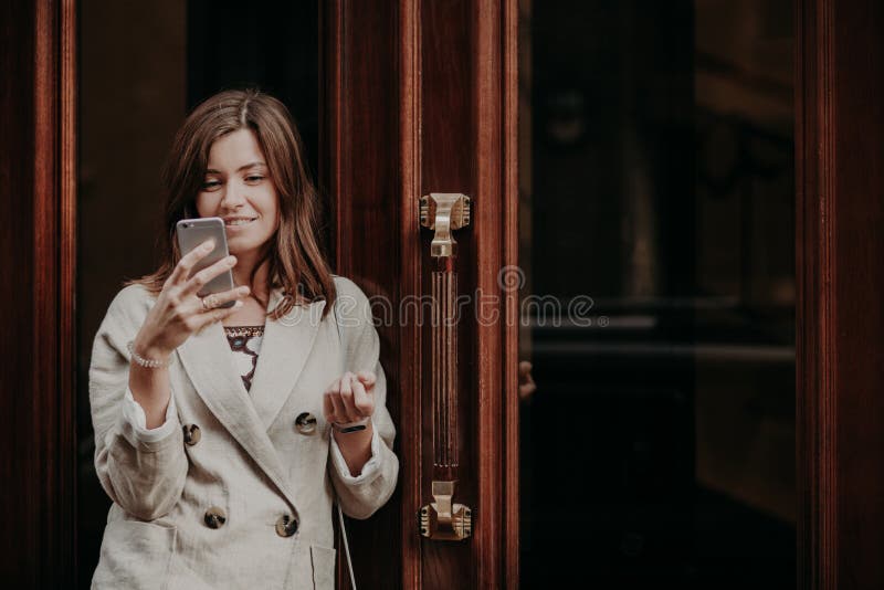 De positieve gelukkige opgetogen Europese vrouw gebruikt online het beleggen op mobiele telefoon, praatjes, gekleed in wit jasje, stock afbeelding De positieve gelukkige opgetogen Europese vrouw gebruikt online het beleggen op mobiele telefoon, praatjes, gekleed in wit jasje, stock afbeelding