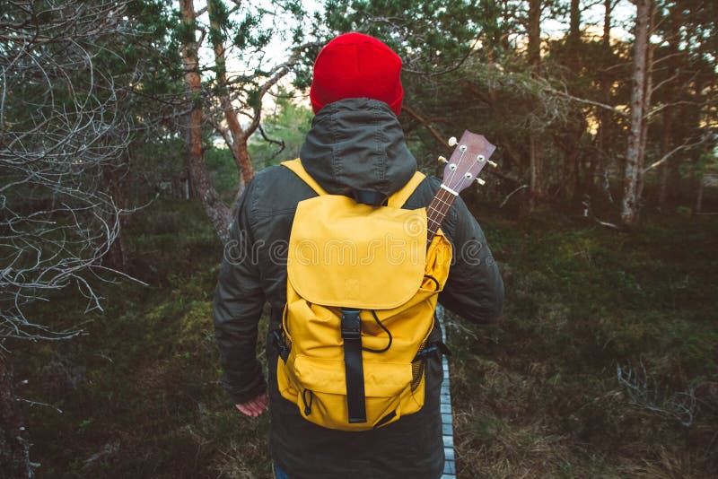 Traveler man is standing on a trail in the middle of a forest with a guitar. Wearing a yellow backpack in a red hat. Lifestyle Travel Concept. Shoot from the back. Traveler man is standing on a trail in the middle of a forest with a guitar. Wearing a yellow backpack in a red hat. Lifestyle Travel Concept. Shoot from the back.