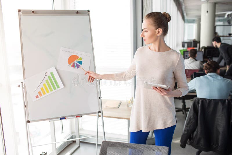De slimme en aardige vrouw bevindt zich dichtbij flipchart en stelt diagrammen op het voor Zij bevindt zich dichtbij groot en hel royalty-vrije stock afbeelding De slimme en aardige vrouw bevindt zich dichtbij flipchart en stelt diagrammen op het voor Zij bevindt zich dichtbij groot en hel royalty-vrije stock afbeelding