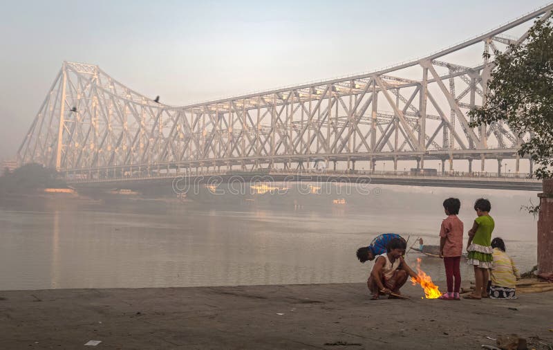 De straatjonge geitjes houden op een koude mistige de winterochtend warm in Mallick ghat dichtbij de brug van Howrah bij de bank stock afbeelding De straatjonge geitjes houden op een koude mistige de winterochtend warm in Mallick ghat dichtbij de brug van Howrah bij de bank stock afbeelding