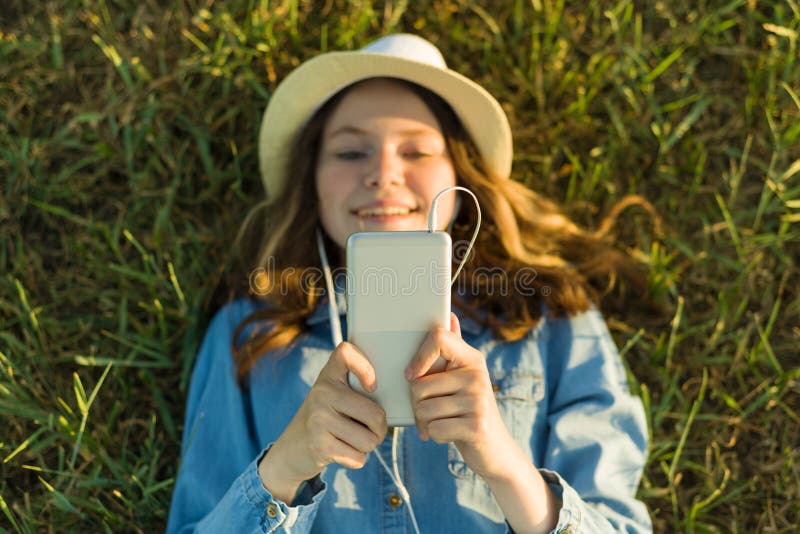 De tiener in hoed met hoofdtelefoons ligt op het groene gras en onderzoekt de telefoon Nadruk op smartphone, hoogste mening royalty-vrije stock afbeeldingen De tiener in hoed met hoofdtelefoons ligt op het groene gras en onderzoekt de telefoon Nadruk op smartphone, hoogste mening royalty-vrije stock afbeeldingen
