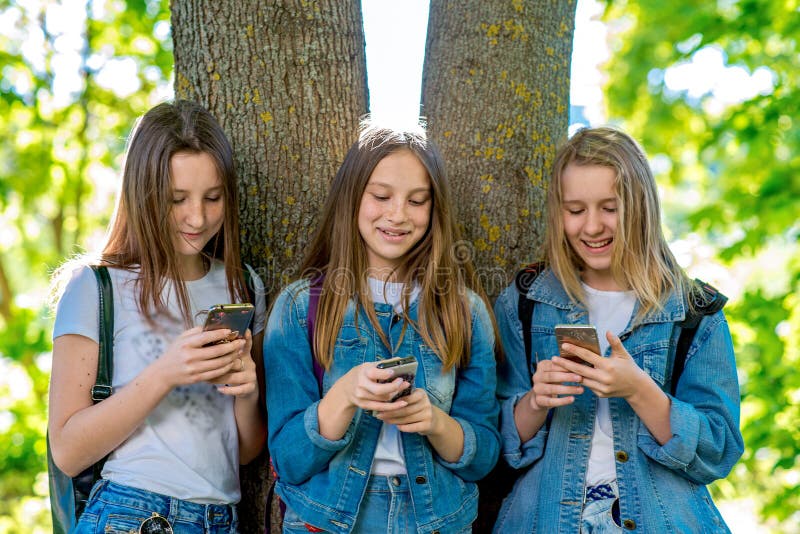 De tiener van het drie meisjesschoolmeisje In de zomerpark door boom Bij de Schoolverandering In handen houdt smartphone De conce stock afbeeldingen De tiener van het drie meisjesschoolmeisje In de zomerpark door boom Bij de Schoolverandering In handen houdt smartphone De conce stock afbeeldingen