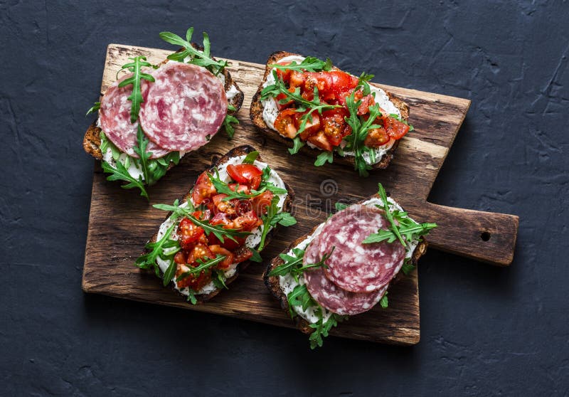De tomaten, de Italiaanse worst en de gehele korrel van de raketsalade paneren bruschetta op een houten hakbord op een donkere ac stock afbeelding De tomaten, de Italiaanse worst en de gehele korrel van de raketsalade paneren bruschetta op een houten hakbord op een donkere ac stock afbeelding