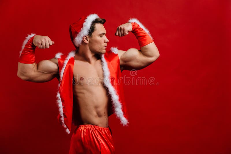 De vakantie van Kerstmis de sexy sterke Kerstman die hoed dragen Jonge spiermens Rode achtergrond stock afbeelding De vakantie van Kerstmis de sexy sterke Kerstman die hoed dragen Jonge spiermens Rode achtergrond stock afbeelding