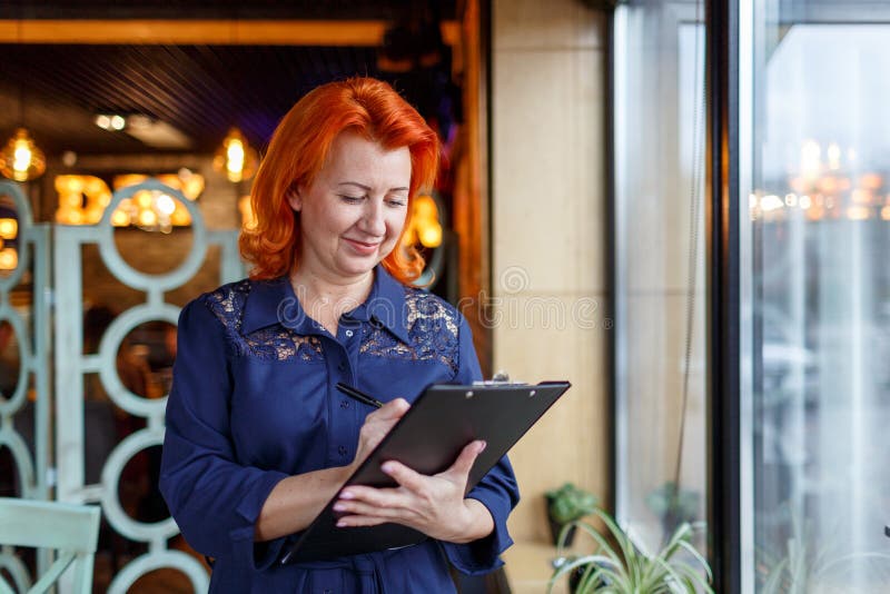 De volwassen vrouw met rood haar, houdt een klembord en schrijft iets stock fotografie De volwassen vrouw met rood haar, houdt een klembord en schrijft iets stock fotografie