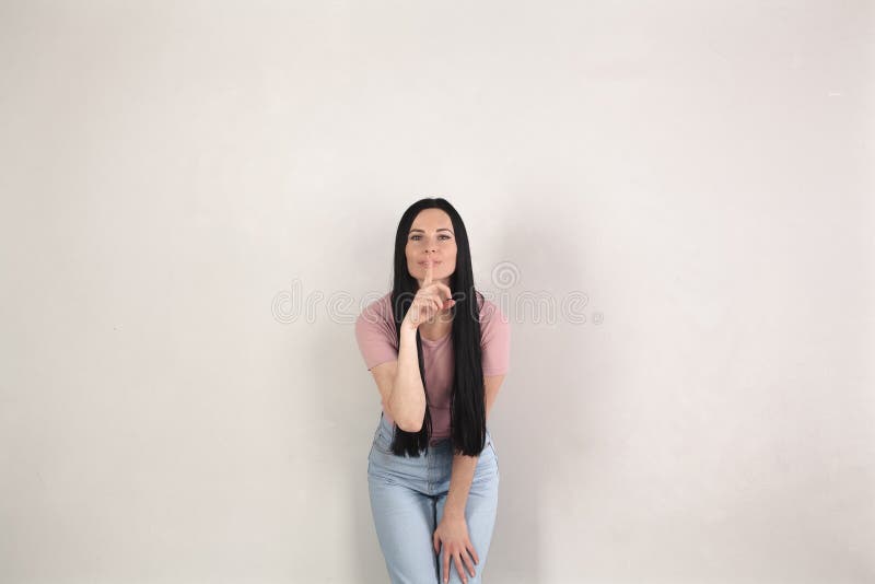 De vrij jonge donkerbruine vrouw met lang haar bevindt zich door de grijze achtergrond die vooruit en stilteteken met het buigen stock fotografie De vrij jonge donkerbruine vrouw met lang haar bevindt zich door de grijze achtergrond die vooruit en stilteteken met het buigen stock fotografie