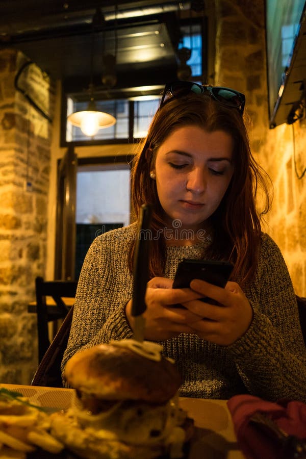 De vrouw babbelt op mobiele telefoon, terwijl in koffiebar zit Het rode hoofd jonge meisje verzendt bericht in smartphone met zon royalty-vrije stock afbeeldingen De vrouw babbelt op mobiele telefoon, terwijl in koffiebar zit Het rode hoofd jonge meisje verzendt bericht in smartphone met zon royalty-vrije stock afbeeldingen