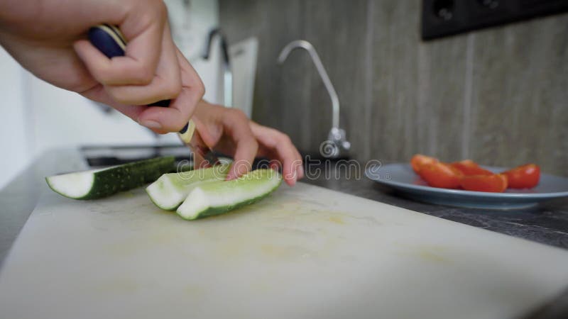 De vrouw snijdt verse komkommers op hakbord op keukenlijst en zettend het in plaat, close-up van haar handen