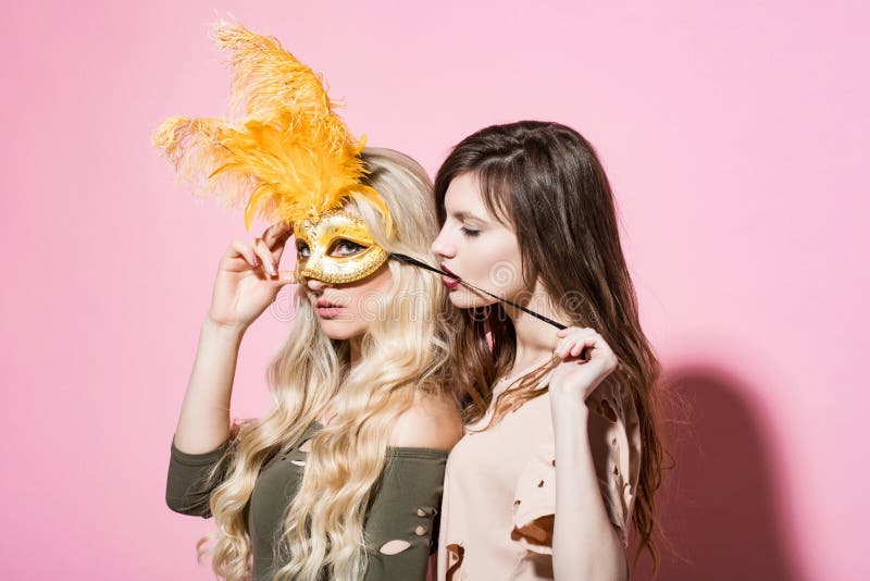 Woman put golden mask on girlfriend face. Love, lgbt, lesbian, lifestyle, romance. Fashion, beauty, look concept. Carnival, party, holidays celebration Girls with long hair on pink background. Woman put golden mask on girlfriend face. Love, lgbt, lesbian, lifestyle, romance. Fashion, beauty, look concept. Carnival, party, holidays celebration Girls with long hair on pink background