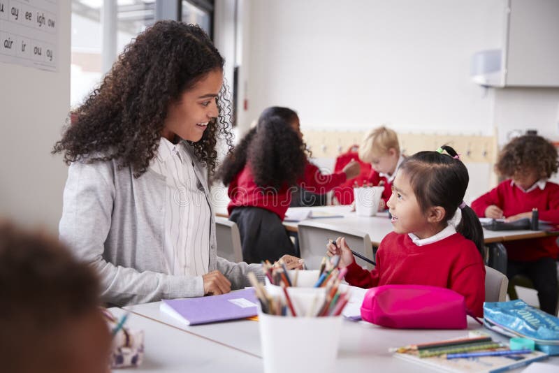 De vrouwelijke zitting van de kleuterschoolleraar bij lijst in een klaslokaal die aan een jong Chinees schoolmeisje spreken, slui royalty-vrije stock afbeeldingen De vrouwelijke zitting van de kleuterschoolleraar bij lijst in een klaslokaal die aan een jong Chinees schoolmeisje spreken, slui royalty-vrije stock afbeeldingen