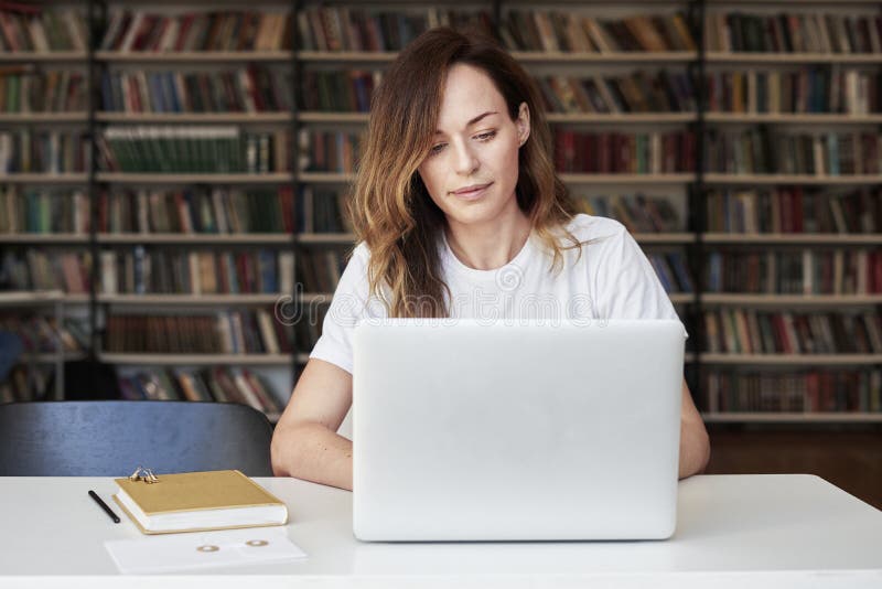 De vrouwenondernemer die aan laptop op mede-werkt kantoor of bibliotheek werken, kijkt slim, boekenrekken Kennis en zelfontplooii
