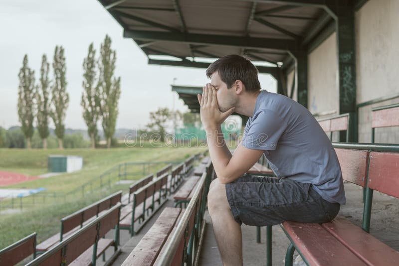 De wanhoop en de gedeprimeerde mens zitten op bank bij stadion stock afbeelding De wanhoop en de gedeprimeerde mens zitten op bank bij stadion stock afbeelding